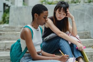 Two student sitting on a sidewalk looking at a notebook.